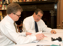 Two men in shirtsleeves work at a table with papers in front of them.