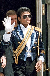 A man wearing dark sunglasses and a jacket covered in blue and yellow rhinestones, holding up his right hand which is covered in a white glove. Behind him stands a man in a black suit.