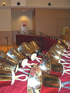 Zdjęcie przedstawia kilkadziesiąt dzwonków ręcznych. Dzwonki położone są obok siebie na długim, zakrzywionym stole. Na stole położony jest czerwony materiał. Przed stołem ustawione są stojaki do nut.