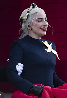 Side view of a smiling Lady Gaga, as she is looking away from the camera, wearing a navy blue dress decorated with a golden bird.
