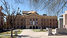 frontal view of the Arizona State Capitol, in winter, framed by the bare limbs of trees, showing the Arizona granite of the building topped by a copper dome
