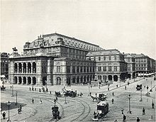 An imposing, heavily ornamented building in a city location, with numerous horsedrawn vehicles and pedestrians passing. There are visible tramlines in the street.