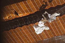 Two black and orange solar arrays, shown uneven and with a large tear visible. A crew member in a spacesuit, attached to the end of a robotic arm, holds a latticework between two solar sails.