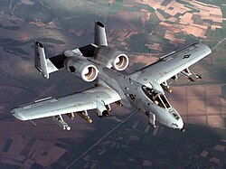 Aerial top view of gray jet aircraft flying above green and brown patchy earth surface. Under each wing are eleven hardpoints for various weapons and ordnance. The two engines are located aft of the wings and in front of two fin units.