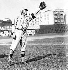 A black-and-white image of a man in a white baseball uniform with a large block "P" on the left breast extends his left arm with a baseball glove on his hand