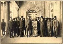 Group shot of dozens of people assembled at the entrance of an imposing building; two columns in view. All subjects face the camera. All but two are dressed in lounge suits: a woman at front-center wears light-coloured Persian garb; the man to her left, first row, wears a white beard and dark-coloured oriental cap and robes.