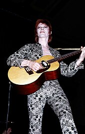 A color photograph of David Bowie with an acoustic guitar