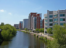 The canalised River Aire flows from the Dark Arches under Leeds's main railway station towards the bottom of the picture. To the left of the river is the lock which links the river with the Leeds and Liverpool Canal. To the right is a riverside walk beneath modern buildings, and in the distance, beyond the railway viaduct and station, are more high-rise modern buildings located on the west side of the city centre.
