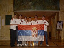 Six boys, standing on a line, all wearing white tops with red logos on their chest. They are holding a red, blue and white striped flag, which features a prominent crown and coat of arms.