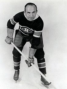 Full profile of a balding ice hockey player in full uniform leaning forward on his stick with a serious look on his face.