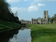 A photograph of a ruined abbey; a river passes by in the lower left hand of the picture, overhung with dark trees. A ruined abbey building in stone makes up the midground of the right side of the photograph.