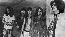 Five smiling men in a row, diagonal to camera angle. The man on the left (farthest to the back) has very long hair and a full beard; he wears a white T-shirt and tie-dyed pants. Next to him, Dave Davies, also with very long hair, wears reflective sunglasses, a black short-sleeved shirt and jeans. In the middle, Mick Avory wears an unbuttoned leather vest and white pants. The man to his right wears a heavy, probably brown leather jacket with a design that is possibly Native American. On the far right, in front, Ray Davies wears a giant paisley kerchief knotted like a tie, over a white jacket.