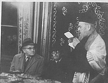 A standing Rabbi Shmuel Benjamin Bachar speaking to David Ben-Gurion and another man, who are seated at a table