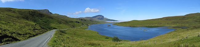 A blue body of water sits beneath a blue sky surrounded by green moorland. A road to the left travels along the lakeside leading towards a small patch of mist and some low hills in the distance.