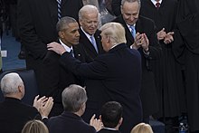 Donald Trump placing his arm on Obama's shoulder. A group of men are clapping in the background and foreground.