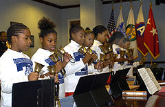 Zdjęcie przedstawia siedem czarnoskórych dziewczynek grających na dzwonkach ręcznych. Dziewczynki stoją przed pulpitami do nut i ubrane są w białe bluzy z logiem szkoły podstawowej. Każda z nich trzyma w każdej dłoni po jednym dzwonku. W tle widocznych jest pięć zawieszonych flag. Wśród nich znajduje się flaga Stanów Zjednoczonych.