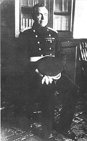 A man seated, wearing a dark uniform with two rows of buttons, with his peaked cap on his lap