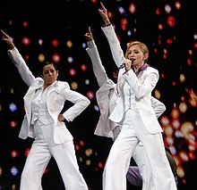 Madonna and her backup singers in white suits, with her right hand stretched up.