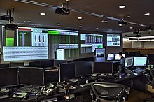 A dark office filled with computers; screens of financial data fill one wall