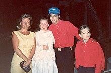 A smiling Jackson wears a blue baseball cap and a red shirt. On his right are two women. One holds a pen, and one holds a small purse. On his left, a young boy looks off-camera. He is dressed in a red shirt, too. Jackson's hand is on his shoulder.