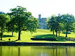 A castle in the distance viewed between trees on a lawn sloping down to a lake in the foreground.