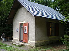 A small grey hut, surrounded by woods, with an open door to which is affixed a picture of the composer