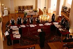 Zdjęcie przedstawia trzynaście osób grających na dzwonkach ręcznych. Dzwonkarze stoją przed stołami ustawionymi w podkowę. Przed nimi stoi dyrygent. Na stołach rozłożone są nuty. Muzycy ubrani są czarne spodnie, białe koszule i czarne kamizelki.