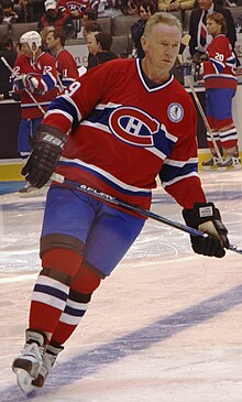 An ice hockey player skating across the ice with a look of concentration on his face. He is in his late fifties, and is wearing a red jersey with blue and white horizontal stripes across the chest, with an H inside of a C logo.