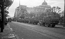 Trucks travelling along wide tree-lined street with large old building in the background
