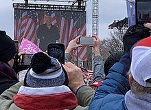 People holding their phones up to take photos and videos of Donald Trump delivering a speech. Trump is not directly visible, but rather a large screen behind the stage shows him.