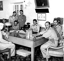 Eight men in dressed in military uniform, posing in a room around a rectangular table. All the men, except for third and fifth persons from the left are seated. The third and fifth person from the left are standing.
