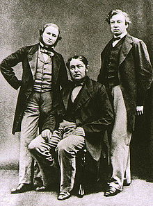 Три средовековни мажи, еден во средината седнува. Сите носат долги јакни, а пократкиот човек лево има брада.