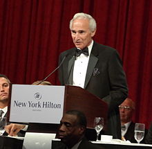 "A elderly man in formal wear makes a speech at a podium during a dinner party."