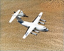 Top view of cargo aircraft in-flight, trailed by a fighter chase aircraft. Under each un-swept wing are two engines suspended forward ahead the leading edge.