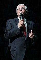 A man in his 50s with white hair speaks into a microphone. He wears a gray suit, red tie, and glasses.
