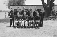 Ten naval officers in uniform sitting for a group portrait: four sitting on a bench, with the other six standing behind. A large, flat-roofed building, two trees, and some scattered people can be seen in the background