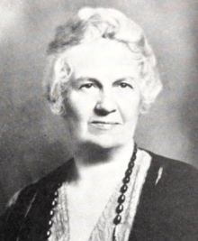 An older white woman with short wavy white hair, wearing dark beads, a print dress, and a dark jacket or cardigan