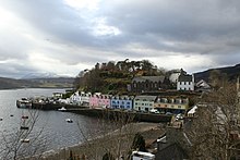 A small harbour fronted with a row of cottages painted in white, pink, green and blue with a tree-covered hillock behind them.