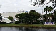 A multi-story building in front of an artificial lake with satellite dishes and communications equipment visible outside. A sign contains the First Coast News logo and logos for WTLV and WJXX.