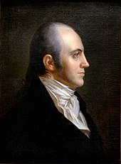 A man with receding gray hair tied in a braid, wearing a high-collared white shirt and black jacket