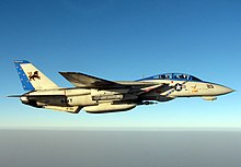 Pale gray jet aircraft flying over water towards right, perpendicular to the camera. Horizon located two-thirds down the photo. Sky made up of two shades, dark blue covers the top, blending with a lighter shade until it is almost white above horizon