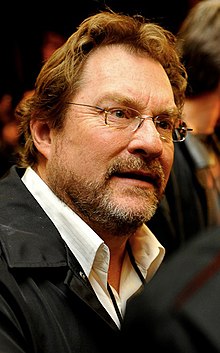 A lightly bearded and bespectacled brown-haired middle-aged Caucasian man wearing a jacket and white shirt with an open collar looks to the camera's right