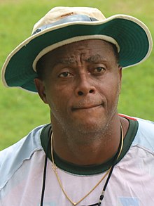 A black man, in a floppy white cricket sun hat, looking ahead, wearing a white training top and a gold-coloured necklace