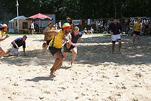 Two teams of players, one in yellow the other in blue, play a form of rugby on a sunlit beach; the central yellow player runs forward, towards the right of picture, clutching the ball with one hand, close to his chest.