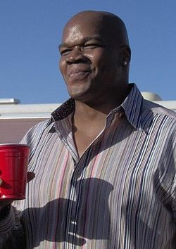 A bald black man in a vertically striped shirt, facing left, smiling and holding a red cup