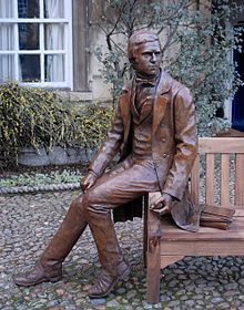 Bronze statue of Darwin in 1830 clothes, seated on the arm of a wooden bench, behind him plants partly cover a stone wall, a window has white-painted wooden frames.