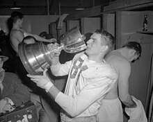 A man with short hair in a white football uniform drinks out of a large silver cup.