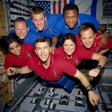 Seven crew members, in red or blue collared shirts, floating in microgravity.