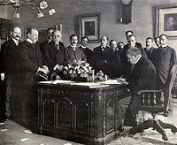 A black and white image of a group of men surrounding the Resolute desk, which had a large bouquet of flowers on it, as Jules Cambon signs the treaty on the desk.