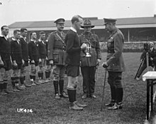 A black and white photo of a rugby field in which three men in military uniform, one of whom is King George V, present a silver trophy to a rugby player dressed in black kit. Behind in a line are the rest of the team.